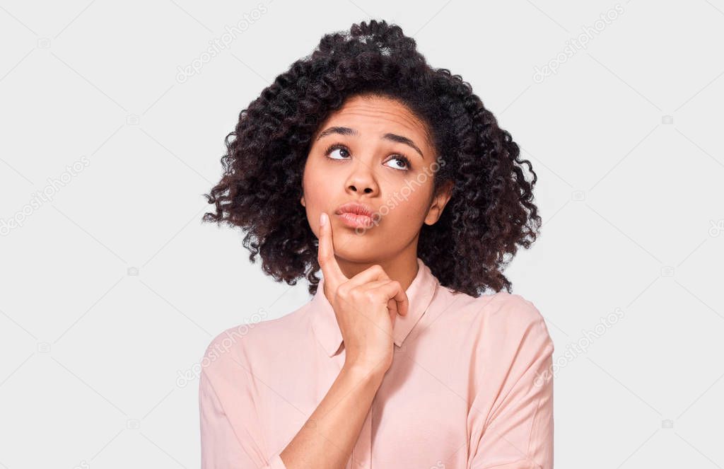 Thoughtful African American woman has puzzled expression, frowns her face and looking at one side with hand on chin. Dark skinned female can`t make choice, has questioned expression, isolated on white
