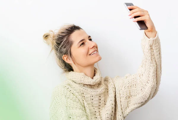 Inomhus bild av leende ung blond kvinna klädd stickad tröja med hår bulle gör Selfie med sin smarta telefon. Kaukasiska kvinnliga innehav mobiltelefon tar självporträtt. — Stockfoto
