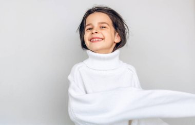Mutlu küçük kız portresi geniş gülümseyen ve büyük beyaz örme kazak dışarı peeks, bir tatil stüdyo açık gri stüdyo duvar üzerinde poz. Güzel çocuk Noel bekliyor