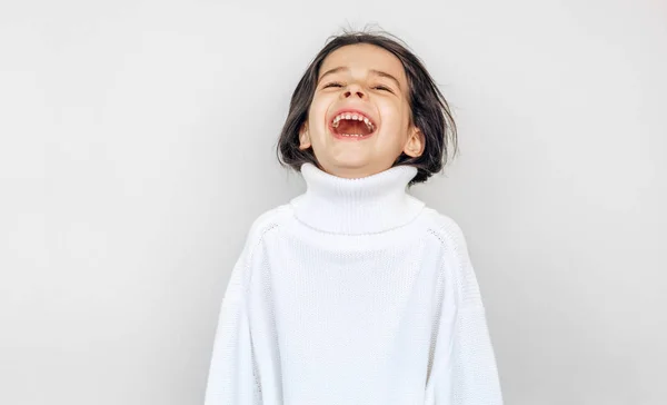 Menina feliz rindo e espreita fora de camisola de malha branca grande, posando sobre estúdio parede estúdio cinza claro em um feriado. Bela criança à espera de presentes de Natal . — Fotografia de Stock