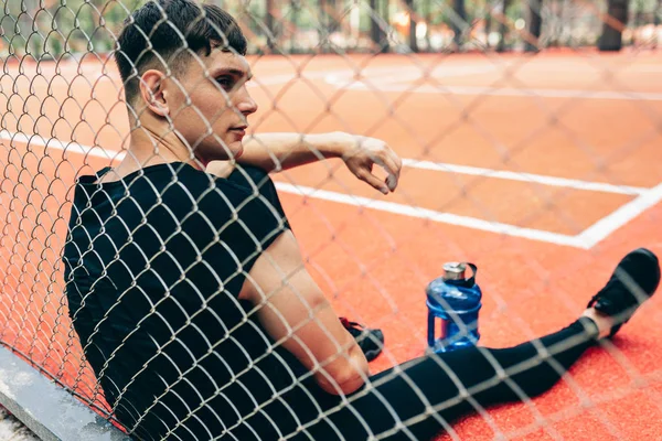 Visão traseira de belo jovem homem fitness descansando após o treino, com uma garrafa azul de água, sentado no sportsground. Saudável masculino fazendo uma pausa depois de se exercitar ao ar livre . — Fotografia de Stock