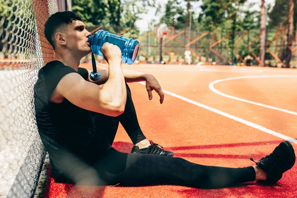 Imagem de visão lateral do jovem atleta descansando após o treino, bebendo água de uma garrafa azul, sentado no sportsground. Saudável masculino fazendo uma pausa depois de se exercitar ao ar livre . — Fotografia de Stock