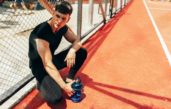 Image of handsome fit young man resting after workout, with a bottle of water on black background. Healthy male taking a break after exercising outdoors. — Stock Photo, Image