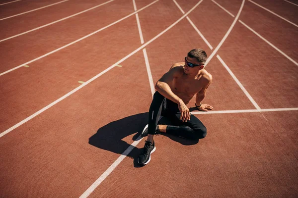 Top uitzicht van de jonge atleet man zittend op race track in stadion nemen rust. Professionele shirtless sportman rusten na de training sessie. Mensen, sport en gezonde levensstijl — Stockfoto