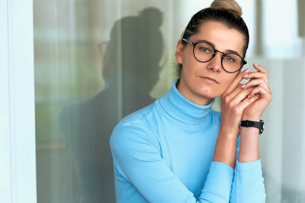 Imagem interior horizontal de bela loira jovem mulher séria no escritório vestindo gola alta azul e óculos, mãos dobradas ao lado de seu rosto e expressão confiante. Pessoas e conceito de negócio — Fotografia de Stock