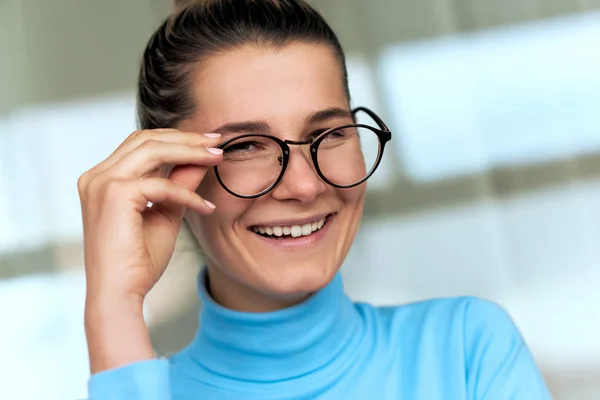 Interior close-up retrato de mulher bonita sorridente no escritório olhando através de óculos da moda, vestindo gola alta azul. Pessoas e empresas — Fotografia de Stock