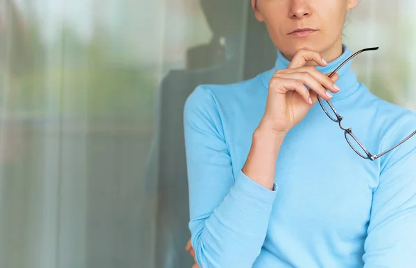 Horizontal recortada imagem de bela loira jovem mulher séria no escritório vestindo gola alta azul, segurando óculos na mão. Pessoas e conceito de negócio — Fotografia de Stock
