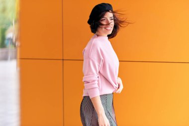 Şehirde sıcak hava zevk rüzgarlı saçlı güzel esmer kadın. Modern şık kadın şehir caddesinde turuncu duvara karşı pembe kazak ve siyah şapka bere giyiyor. Doğal aydınlatma