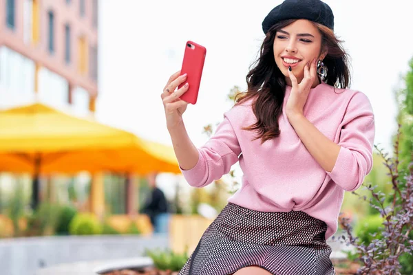 Городской портрет великолепной молодой женщины, сидящей на улице, делающей автопортрет на смартфоне. Брюнетка в розовом свитере, черный берет делает селфи на городской улице . — стоковое фото
