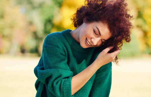 Feliz bela jovem sorrindo com a mão no cabelo, contra o fundo da natureza com cabelo encaracolado ventoso, tem expressão positiva, vestida com suéter verde. Pessoas, viagens e conceito de estilo de vida . — Fotografia de Stock