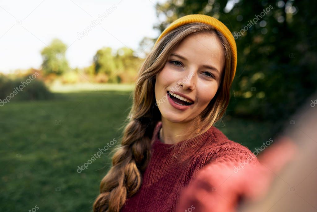 Close-up self portrait of beautiful young blonde woman smiling broadly, wearing sweater and hat, posing on nature background in the park. Happy female taking selfie. People, travel, lifestyle concept.