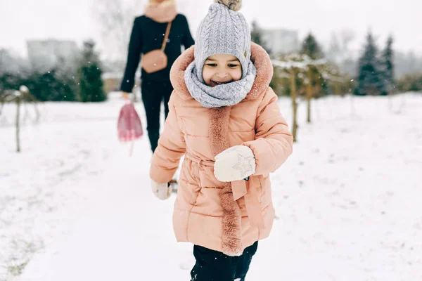 Happy little girl smiling and playing together with her mother in the park on snowing day. Cute child playing catch me with her mom on the snow on winter day. Christmas mood. Childhood