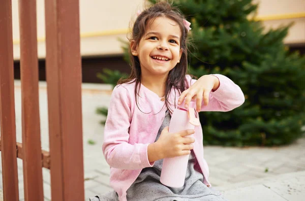 Cute little girl smiling and opening her eco glass bottle for drinking a water, sitting outdoors. Happy child pupil relaxing outside after preschool lessons. People, education concept — 스톡 사진