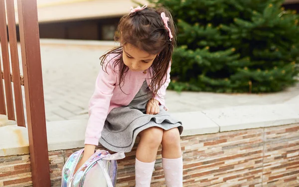 Image of cute little girl preschooler sitting outside and searching something on her backpack. Happy child pupil relaxing, have a break after preschool lessons. People, education concept — Stock Photo, Image