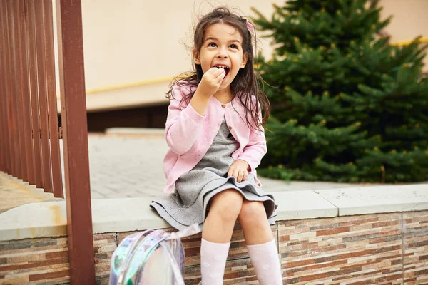 Ao ar livre imagem horizontal da menina bonito pré-escolar sentado do lado de fora e almoçando. Aluno feliz criança relaxante, ter uma pausa após as aulas da escola. Pessoas, conceito de educação — Fotografia de Stock