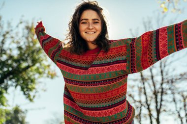 Rüzgarlı saçları ve çilleriyle gülümseyen, güz havasının tadını çıkaran, güneş ışığı arka planında süveter giyen mutlu, güzel, esmer bir kadın. Kolları açık gülümseyen bir kadın..