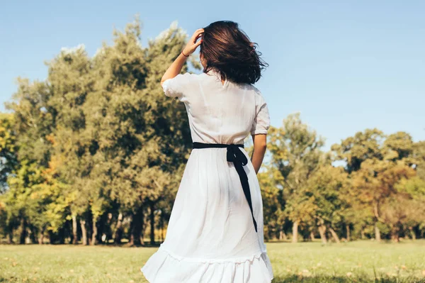 Visão traseira imagem de mulher morena com cabelo ventoso apreciando o clima quente, vestindo vestido branco no fundo da natureza. Jovem bela mulher andando no parque no dia ensolarado . — Fotografia de Stock