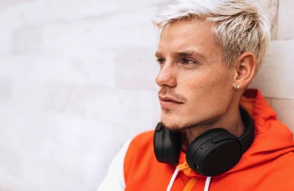 Close-up portrait of handsome young man looking at one side with headphones on the neck, sitting against  light grey outdoor background. Lifestyle, people, technology and music concept. — Stock Photo, Image