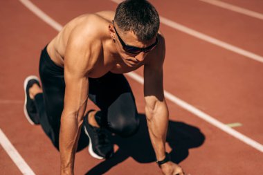Stadyumdaki hipodromda koşmaya hazırlanan üstsüz bir sporcunun yakın plan görüntüsü. Erkek koşucu yarışma için antrenman sırasında koşuyor. İnsanlar, spor ve sağlıklı yaşam tarzı