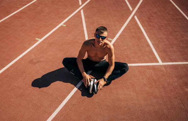 Νεαρός αθλητής που κάθεται στον ιππόδρομο στο στάδιο και ξεκουράζεται. Επαγγελματίας αθλητής χωρίς πουκάμισο ξεκουράζεται μετά την προπόνηση. Άνθρωποι, αθλητισμός και υγιεινός τρόπος ζωής — Φωτογραφία Αρχείου