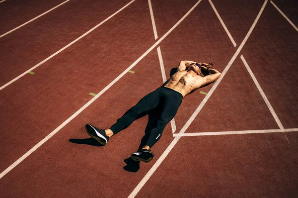 Πάνω άποψη εικόνα του νεαρού αθλητή shirtless ξαπλωμένος σε πίστα στο γήπεδο λαμβάνοντας υπόλοιπο. Επαγγελματίας αθλητής που ξεκουράζεται μετά την προπόνηση. Άνθρωποι, αθλητισμός και υγιεινός τρόπος ζωής — Φωτογραφία Αρχείου