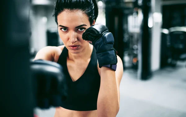 Close-up portrait of young woman boxer hitting a huge punching bag at a boxing studio. Sportswoman kickboxer training hard in the gym. Sport, lifestyle and people concept. — Stock Photo, Image