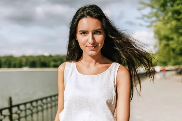 Beautiful young brunette woman smiling broadly with a windy blowing long hair in the park on a background with a lake. — Stock Photo, Image