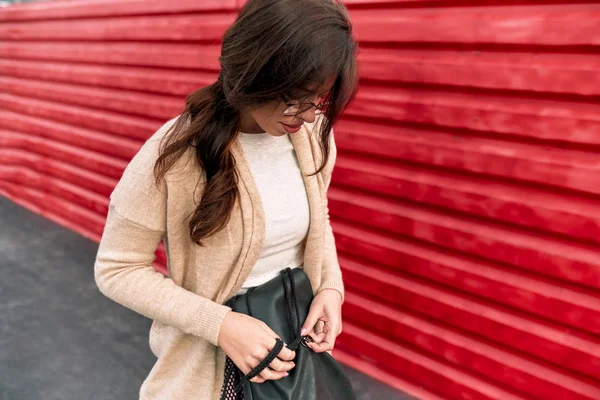 Joven estudiante que busca algo en su mochila, mientras camina por la calle de la ciudad contra la pared roja. Personas y estilo de vida . —  Fotos de Stock