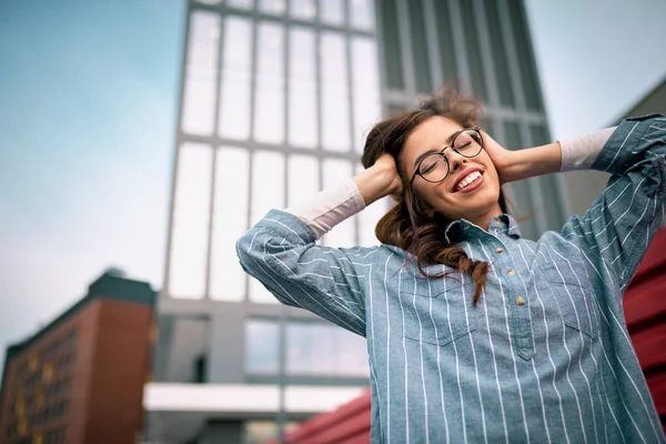 Retrato de metade do comprimento da jovem mulher usando camisa azul de mãos dadas na cabeça enquanto estava perto do prédio de escritórios. Mulher de negócios bem sucedida sorrindo com os olhos fechados ao ar livre . — Fotografia de Stock