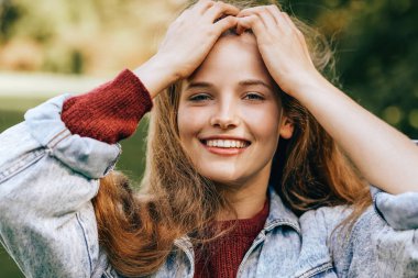 Close-up portrait of beautiful happy young woman smiling broadly with healthy toothy smile, wearing red sweater and denim jacket, posing on nature background in the park. People, travel, lifestyle clipart