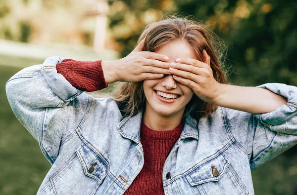 Cheerful beautiful young woman smiling broadly with healthy toothy smile, wearing red sweater and denim jacket, covering her eyes with both hands posing on nature background. People, travel, lifestyle — Stock Photo, Image