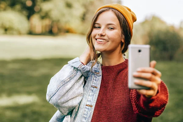 Retrato de bela jovem loira sorrindo amplamente, vestindo suéter vermelho e chapéu amarelo, posando no fundo da natureza no parque. Uma mulher feliz a tirar selfie. Pessoas, viagens, conceito de estilo de vida . — Fotografia de Stock