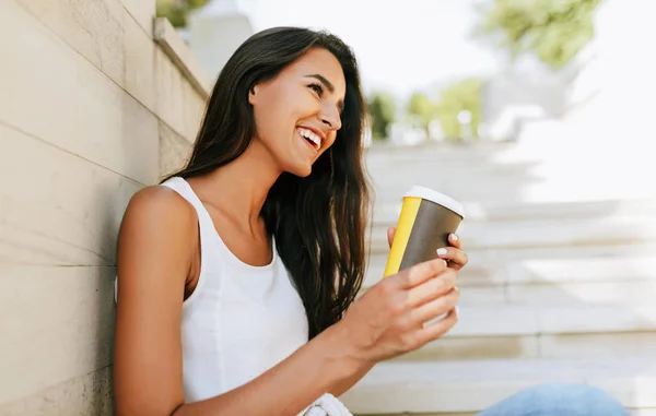 Vista lateral imagem da bela mulher morena sorrindo e segurando xícara descartável de café na rua da cidade. Muito feminino tomar café na mão. Estudante bonita beber café ao ar livre . — Fotografia de Stock