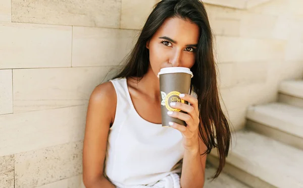 Vakker brunettdame har en kopp kaffe på byens gate. Vakker kvinnelig kaffe i hånden. Pene elever som drikker kaffe utendørs om morgenen . – stockfoto
