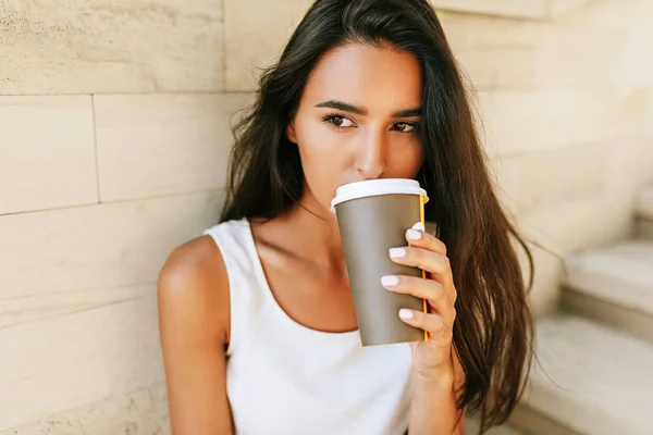 Close-up portrait beautiful brunette woman holds disposable cup of coffee in the city street. Pretty female takeaway coffee in hand. Pretty student drinking coffee outdoors in the morning. — Stock Photo, Image