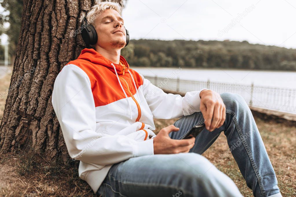 Full body view image of handsome young man enjoying weather in city park, relaxing while listening to his favorite music from wireless headphones in the park next to the tree.