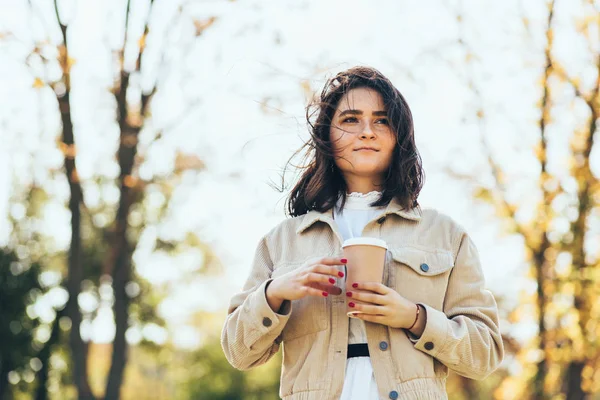 Mulher morena bonita segura xícara descartável de café durante a caminhada no parque. Happy female takeaway café nas mãos. Estudante bonita bebendo café ao ar livre pela manhã . — Fotografia de Stock