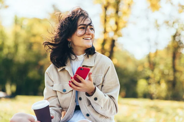 Jovem feliz segura xícara descartável de café, tirando foto no telefone inteligente durante a caminhada no parque. Bela estudante bebendo café takeaway e navegação on-line no telefone móvel . — Fotografia de Stock