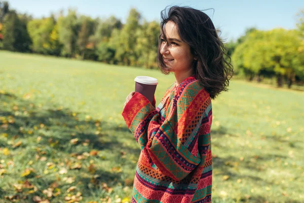 Усміхнена красива молода жінка в светрі і тримає одноразову чашку кави в міському парку. Гарненька студентка п'є каву на відкритому повітрі вранці, йдучи до університету . — стокове фото