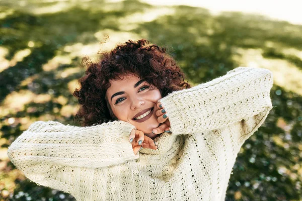Красива молода жінка, яка посміхається з зубною посмішкою, позує на фоні природи з кучерявим волоссям, має позитивний вираз, одягнений в трикотажний светр. Люди, спосіб життя — стокове фото