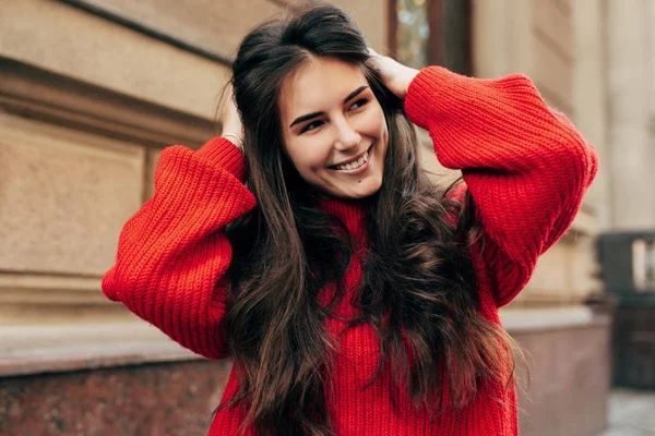 Mulher morena jovem bonita sorrindo amplamente com as mãos em seu cabelo longo. Retrato ao ar livre de modelo feminino bonito em camisola vermelha de malha na moda posando durante a caminhada na rua da cidade . — Fotografia de Stock