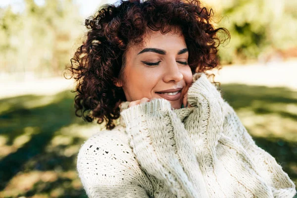 Outdoor close-up portrait of beautiful young woman smiling with closed eyes, posing against nature background with curly hair, have positive expression, wearing knitted sweater. People, lifestyle