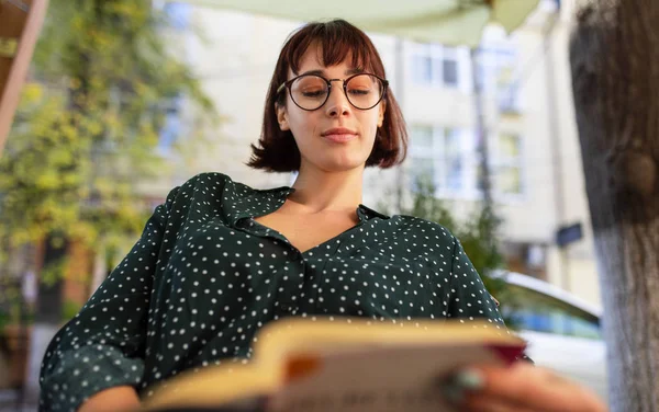 Imagem de bela jovem mulher vestindo camisa verde com pontos brancos e óculos transparentes, sentado ao ar livre na rua da cidade e livro de leitura. Jovem estudante aprendendo na rua da cidade . — Fotografia de Stock