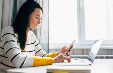 Evde çalışan esmer genç bir kadının yan görüntüsü. Beyaz kadın, evdeki masada dizüstü bilgisayarla oturuyor ve cep telefonundan sosyal medyada yeni yazılar yayınlıyor.