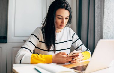 Evden çalışan genç bir kadının kapalı mekan görüntüsü. Beyaz kadın girişimci mutfaktaki masada dizüstü bilgisayarla oturuyor ve cep telefonundaki yeni mesajları kontrol ediyor..