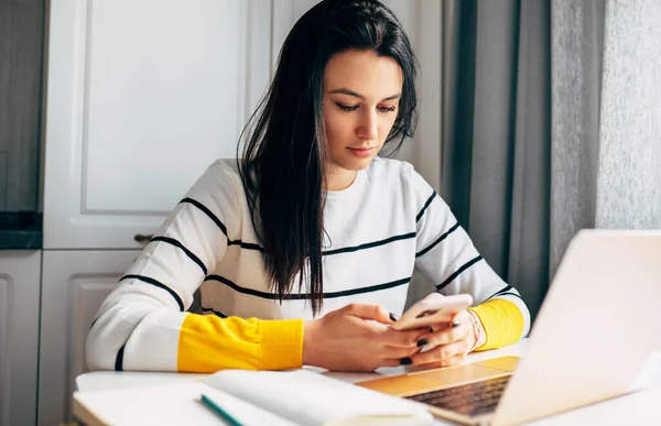自宅から働く若い女性の屋内イメージ 白人女性起業家は ラップトップコンピュータでキッチンに座って 携帯電話上の新しいメッセージをチェックします — ストック写真