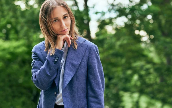 Outdoor portrait of a beautiful female entrepreneur in blue suit looking to the camera and resting in the park after work. Young woman smiling, relaxing, and posing outside.