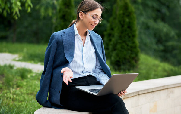Beautiful young woman sitting in the park working on a laptop. Stylish entrepreneur female wearing a trendy outfit is reading an email letter on laptop computer outdoor.