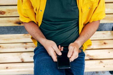 Kapalı çekimde, dışarıda bankta oturan genç adamın cep telefonunu kontrol edişinin görüntüsü. Sarı tişörtlü, yeşil tişörtlü ve kot pantolonlu bir erkek şehrin caddesinde akıllı telefondan online alışveriş yapıyor..