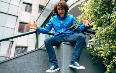 Açık havada mavi yağmurluk giyen, kollarını kaldırmış bir binanın merdivenlerinde oturan neşeli genç adamın görüntüsü. Yağmurlu bir günde bir erkek sokakta dinleniyor. Hippi adamın neşeli duyguları var..